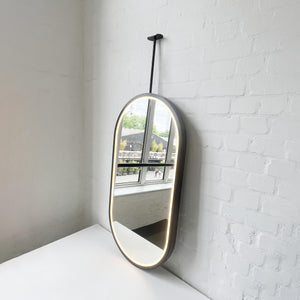 Modern front illuminated Capsula™ ceiling suspended pill shaped mirror with a black powder coated full frame
