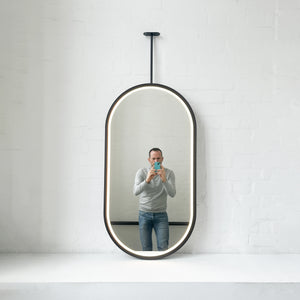 Modern front illuminated Capsula™ ceiling suspended pill shaped mirror with a black powder coated full frame.